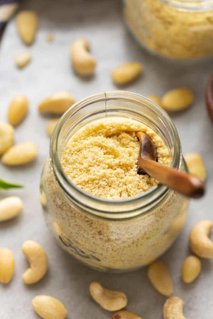 A small jar of Homemade vegan parmesan cheese with a little wooden spoon inside surrounded by cashews and almonds