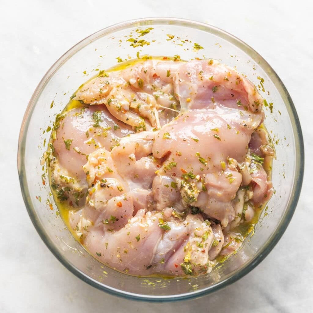 A bowl of boneless, skinless chicken thighs marinating in chimichurri sauce.