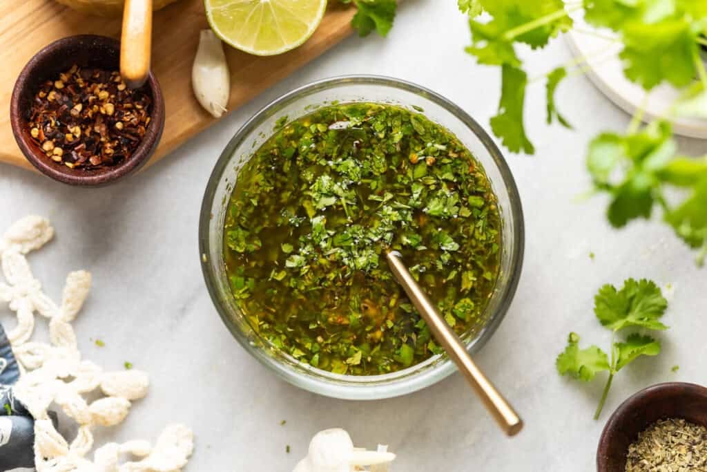 A glass bowl with cilantro chimichurri and a fork with a small bowl of red chili flakes, oregano, a garlic clove, half a lime and fresh cilantro surrounding the bowl.