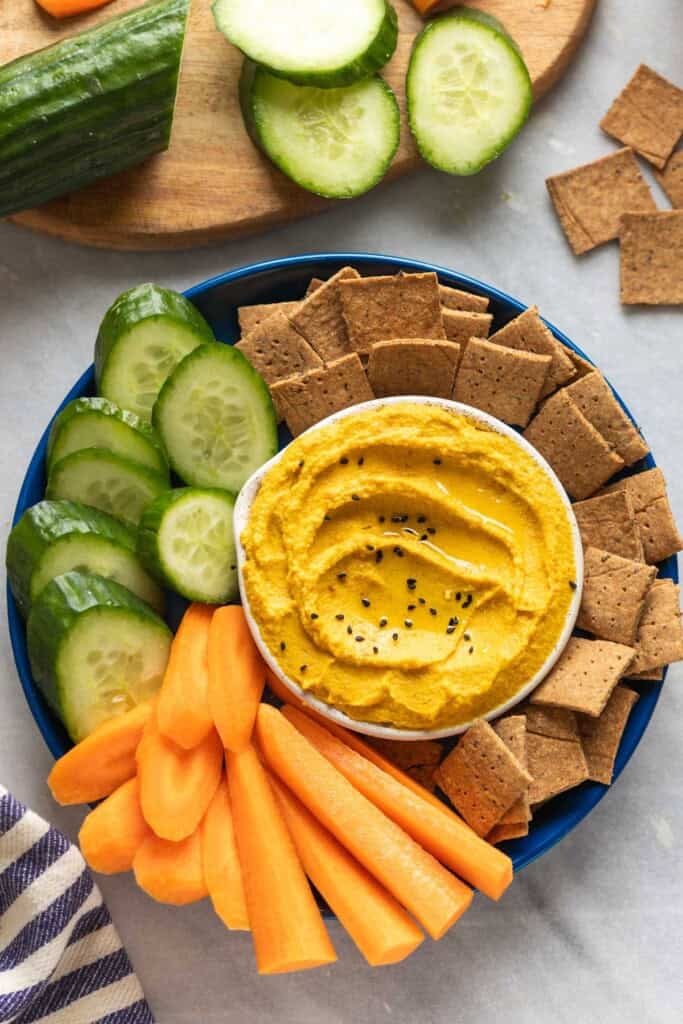 Roasted Carrot Hummus topped with black sesame seeds in a dipping bowl on a plate with sliced carrots, cucumber and homemade almond pulp crackers. With cucumber and crackers on the side