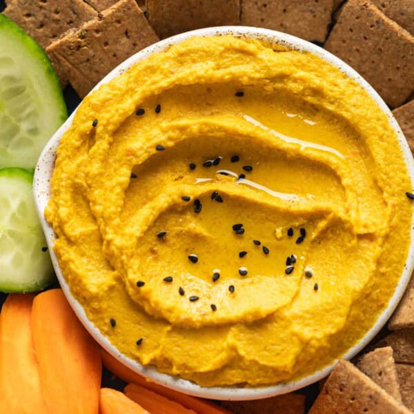 Roasted Carrot Hummus topped with black sesame seeds in a dipping bowl on a plate with sliced carrots, cucumber and homemade almond pulp crackers.