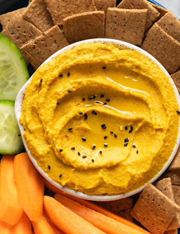 Roasted Carrot Hummus topped with black sesame seeds in a dipping bowl on a plate with sliced carrots, cucumber and homemade almond pulp crackers.