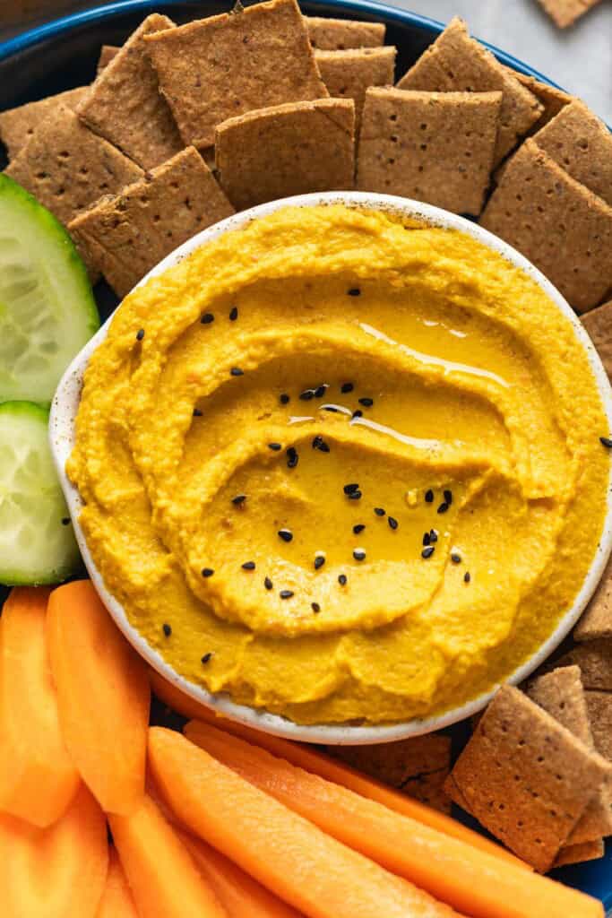 Roasted Carrot Hummus topped with black sesame seeds in a dipping bowl on a plate with sliced carrots, cucumber and homemade almond pulp crackers.