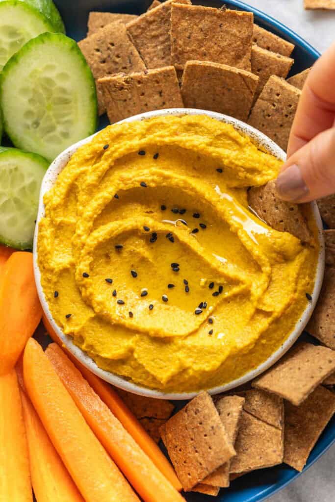 A hand dipping an almond pulp cracker into a bowl of roasted carrot hummus