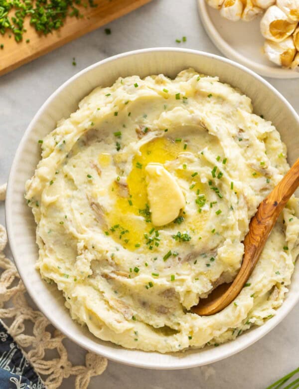 Roasted Garlic Mashed Potatoes in a serving bowl with melted butter and chopped chives on top and a serving spoon in the bowl.