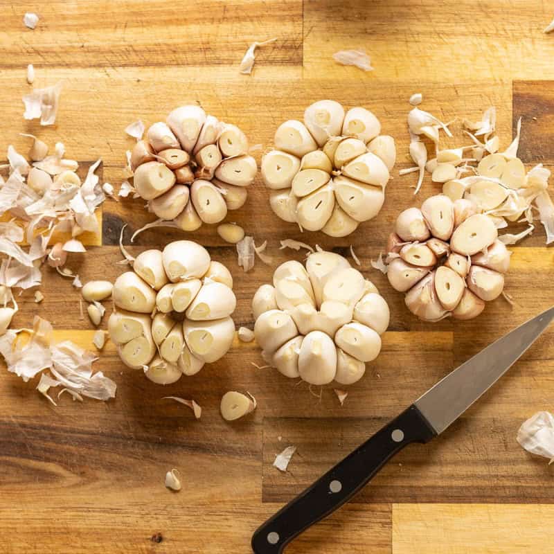 Five raw heads of garlic with the outer most peel removed and the tops cut off on a cutting board.