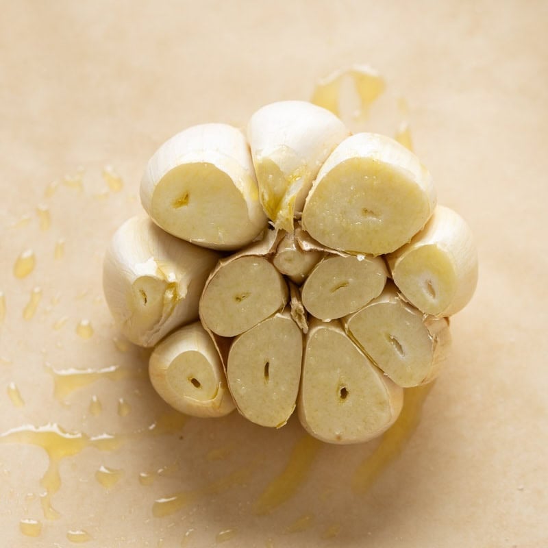 A raw head of garlic with the top cut off and seasoned with olive oil and salt on a piece of parchment paper.
