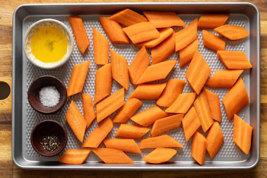 Raw carrots on a baking sheet with little bowls of salt, pepper and oil