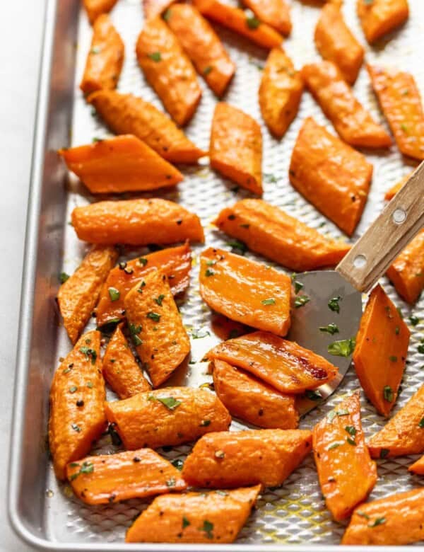 Roasted carrots with fresh chopped parsley on a baking sheet with a small spatula scooping up some of the carrots.