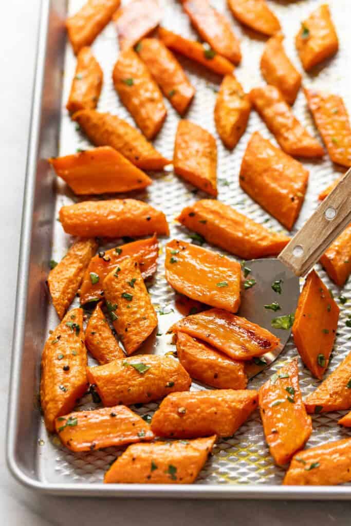 Roasted carrots with fresh chopped parsley on a baking sheet with a small spatula scooping up some of the carrots.