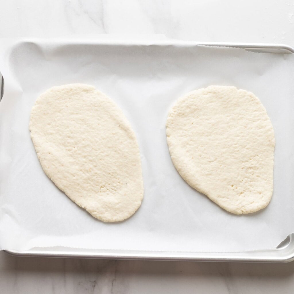 two pieces of gf naan dough rolled out into tear drop shapes, resting on a baking sheet lined with baking paper