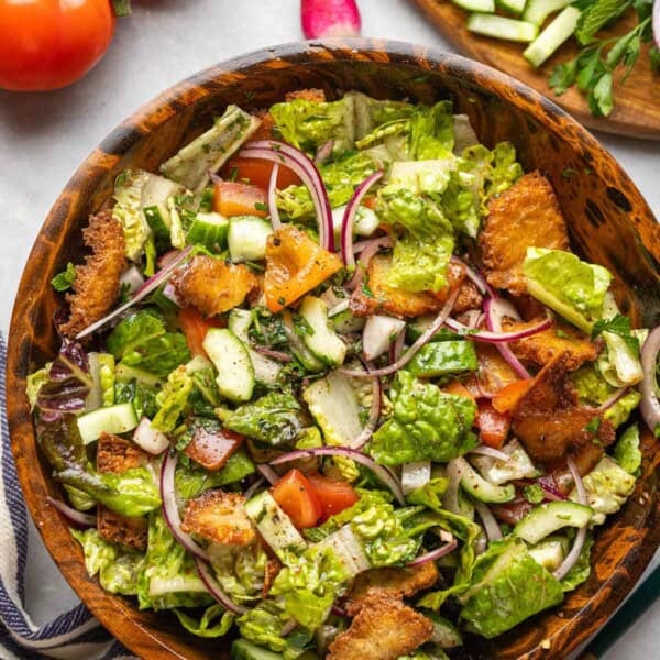 Fattoush salad made with tomatoes, red onion, cucumber, radishes, mint, romaine lettuce and gluten free fried pita chips in a wooden salad bowl surrounded by the fresh ingredients, a kitchen towel and a serving fork & spoon on the side.
