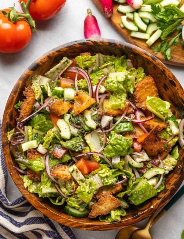 Fattoush salad made with tomatoes, red onion, cucumber, radishes, mint, romaine lettuce and gluten free fried pita chips in a wooden salad bowl surrounded by the fresh ingredients, a kitchen towel and a serving fork & spoon on the side.