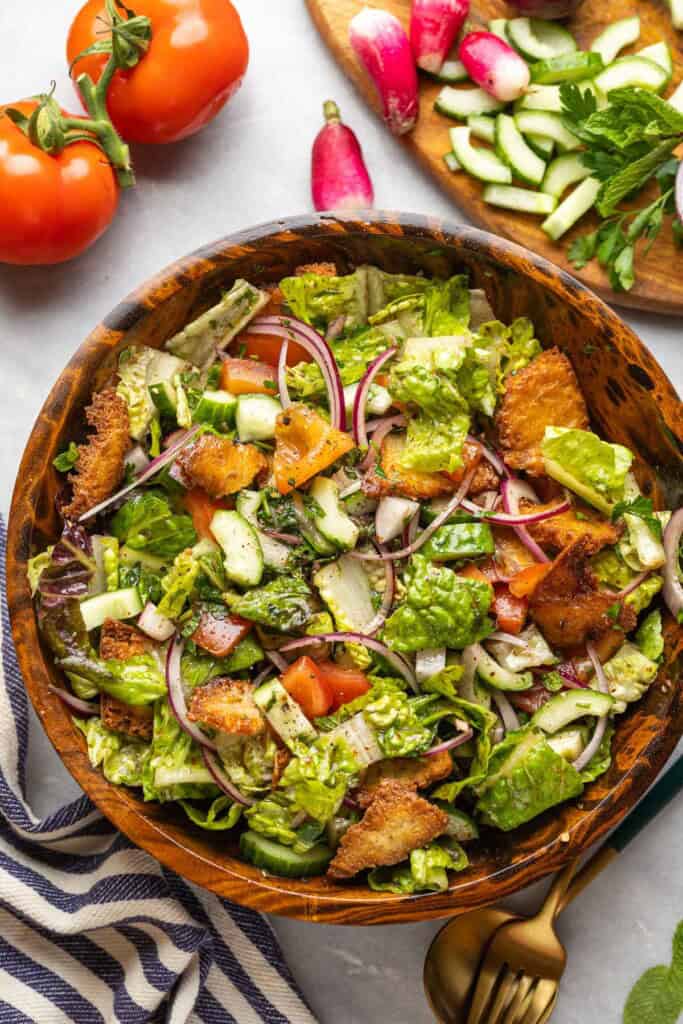 Fattoush salad made with tomatoes, red onion, cucumber, radishes, mint, romaine lettuce and gluten free fried pita chips in a wooden salad bowl surrounded by the fresh ingredients, a kitchen towel and a serving fork & spoon on the side.