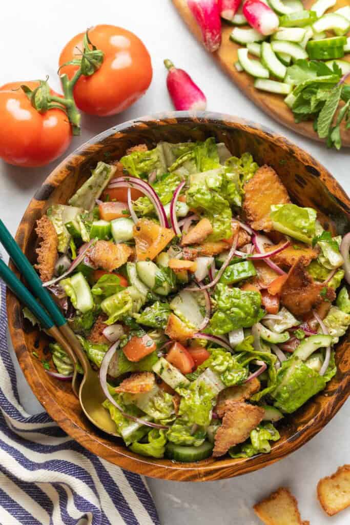 Fattoush salad made with tomatoes, red onion, cucumber, radishes, mint, romaine lettuce and gluten free fried pita chips in a wooden salad bowl with a pair of serving spoons, surrounded by the fresh ingredients.