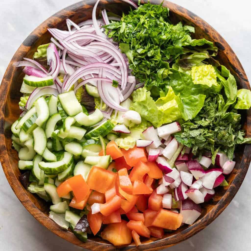 A salad bowl full of unmixed fattoush salad ingredients: tomatoes, red onion, cucumber, radishes, mint and romaine lettuce.