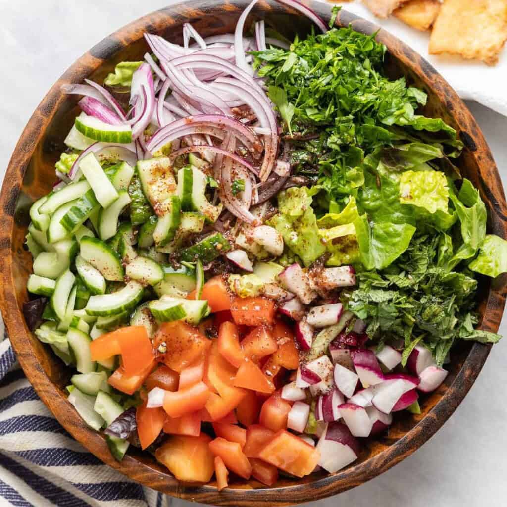 A salad bowl full of unmixed fattoush salad ingredients: tomatoes, red onion, cucumber, radishes, mint and romaine lettuces with the pomegranate vinaigrette poured over the top