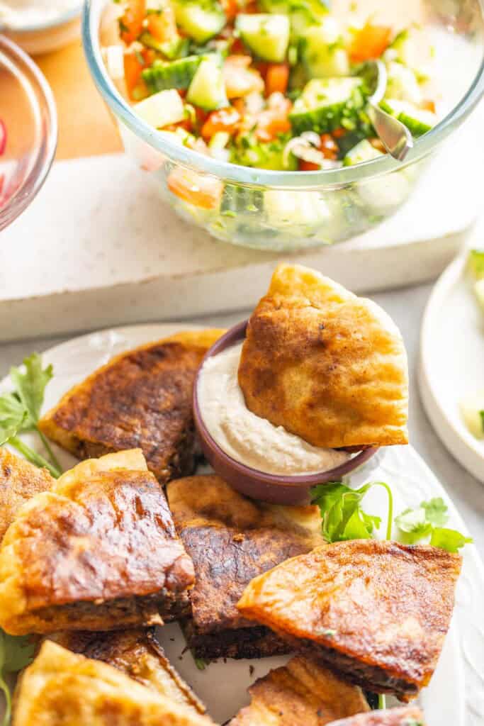 A gluten free arayes wedge dipped into a small bowl of tahini yoghurt sauce with a cucumber & tomato salad on the side