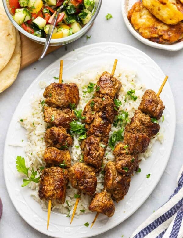 Three skewers of Shawarma Chicken Kebab over a bed of rice with fresh parsley on top, fried halloumi and a cucumber tomato salad on the side.