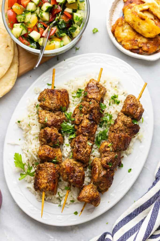 Three skewers of Shawarma Chicken Kebab over a bed of rice with fresh parsley on top, fried halloumi and a cucumber tomato salad on the side.