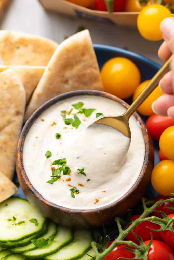 A spoon scooping up some Tahini Yogurt Sauce in a dipping bowl surrounded by pita wedges, cucumber slices and cherry tomatoes.