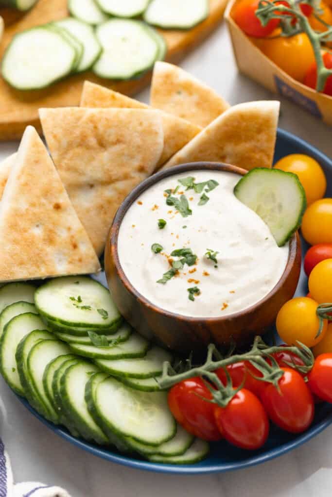 A Tahini Yogurt Sauce in a dipping bowl with a cucumber slice in the sauce and fresh chopped parsley scattered on top in the middle of a plate full of pita wedges, cucumber slices and cherry tomatoes.