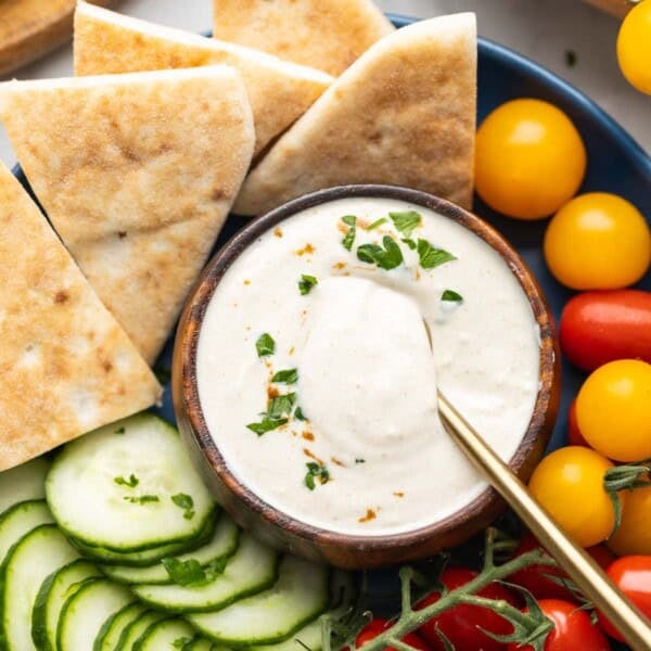 A Tahini Yogurt Sauce in a dipping bowl in the middle of a plate full of pita wedges, cucumber slices and cherry tomatoes.