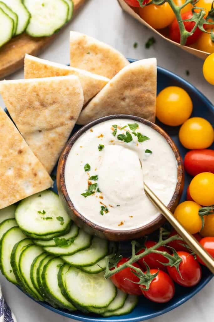 A Tahini Yogurt Sauce in a dipping bowl in the middle of a plate full of pita wedges, cucumber slices and cherry tomatoes.