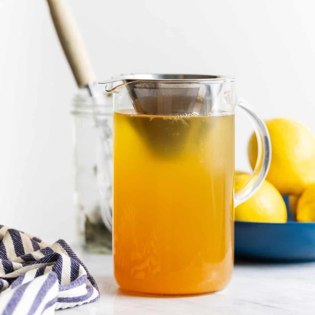 Cold brewed green tea strained into a pitcher with lemonade through a metal cone sieve with a bowl of lemons in the back