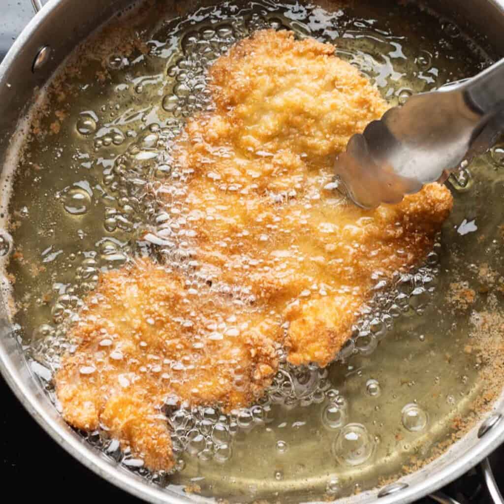 A pair of kitchen tongs lifting up gluten free schnitzel from a pan full of frying oil