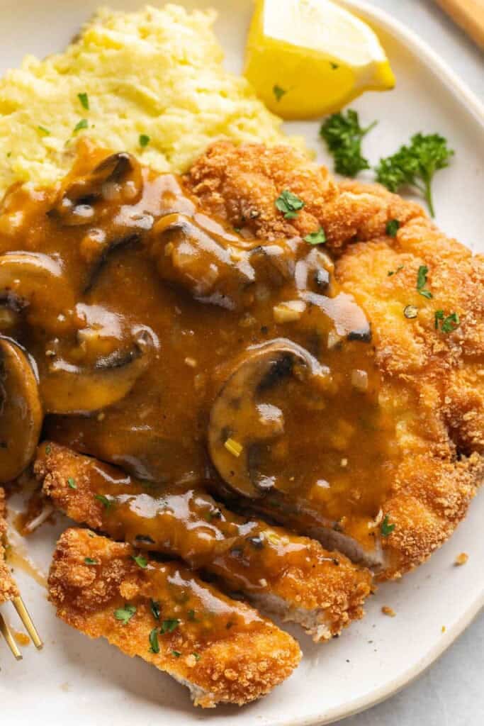 Gluten free schnitzel covered with a mushroom gravy and fresh, chopped parsley with a side of mashed potatoes and a lemon wedge.