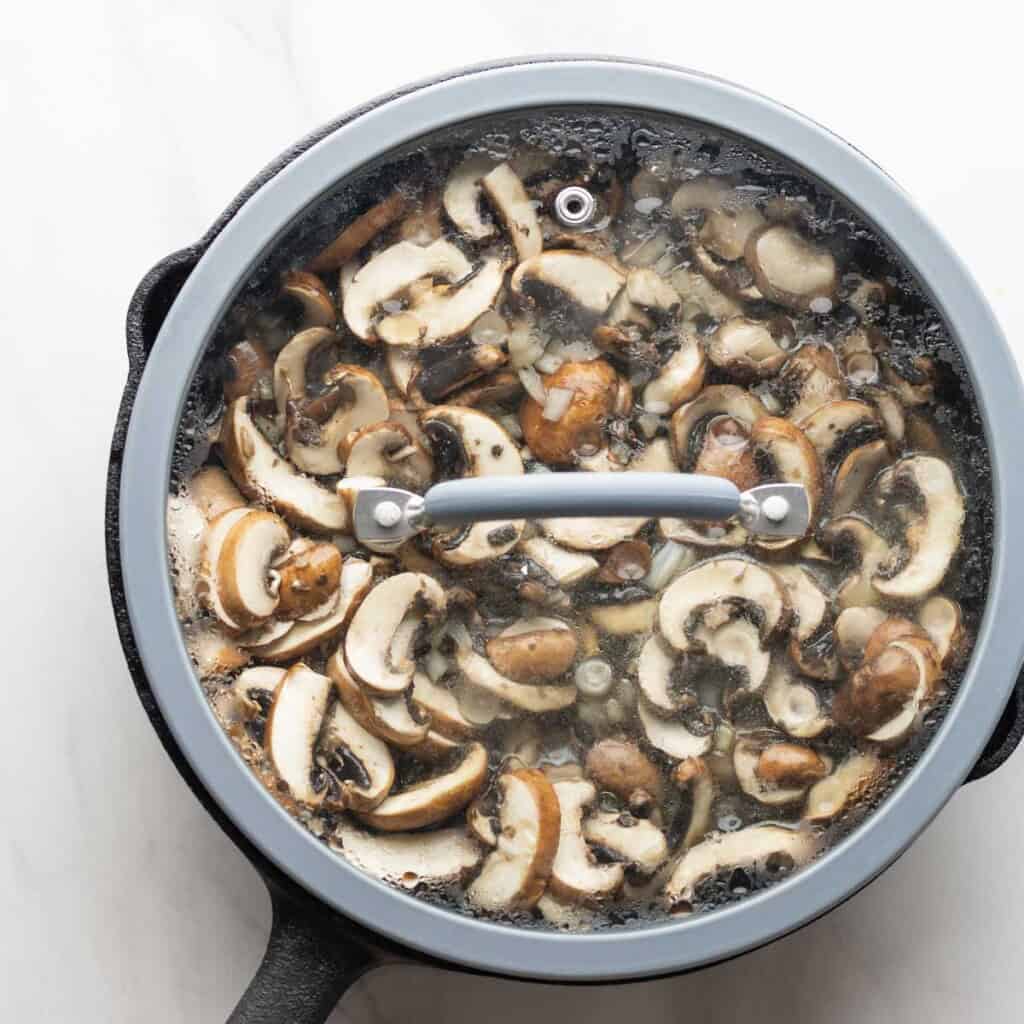 a cast iron pan full of sliced mushrooms and onions with a lid on top