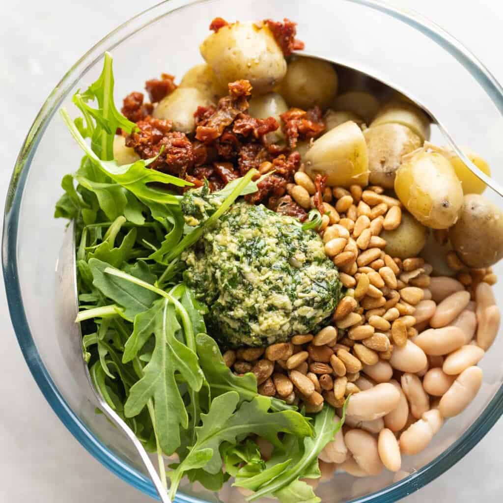 Pesto potato salad ingredients in a bowl: boiled potatoes, cannellini beans, toasted pine nuts, sundried tomatoes, arugula and pesto