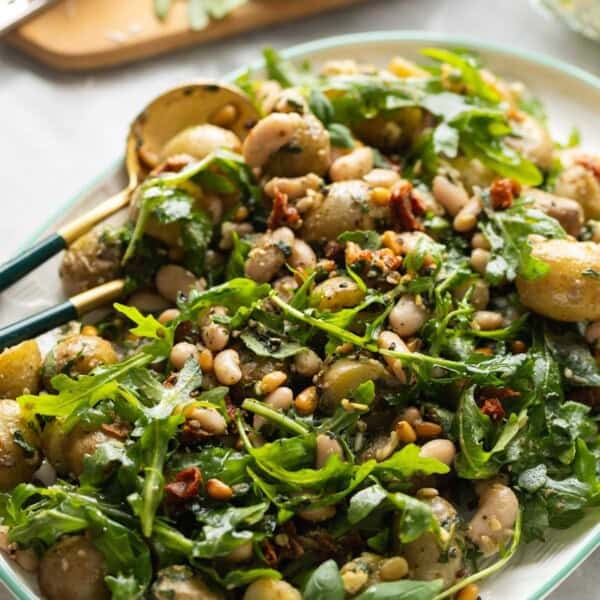 Potato Pesto Salad made with boiled potatoes, white beans, sundried tomatoes, toasted pine nuts, rocket and pesto on a serving platter with serving spoons.