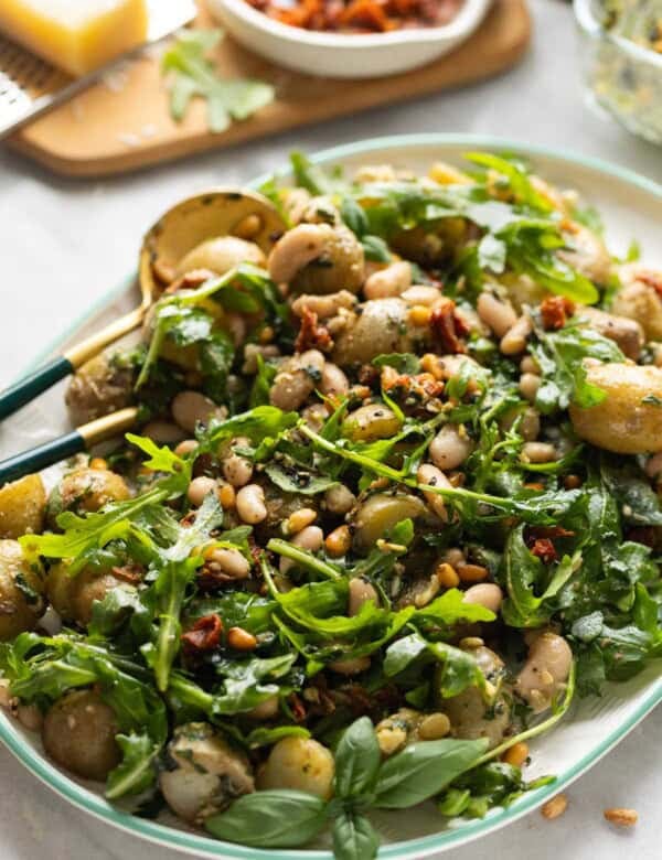 Potato Pesto Salad made with boiled potatoes, white beans, sundried tomatoes, toasted pine nuts, rocket and pesto on a serving platter with serving spoons.