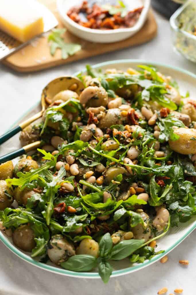 Potato Pesto Salad made with boiled potatoes, white beans, sundried tomatoes, toasted pine nuts, rocket and pesto on a serving platter with serving spoons.