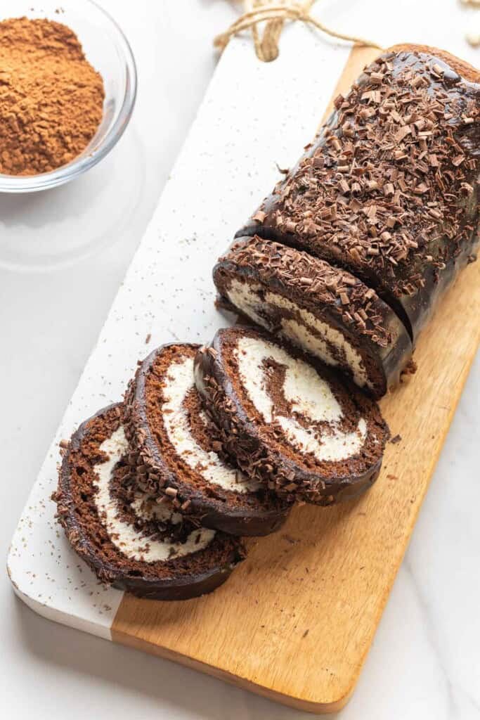 Gluten free chocolate swiss cake roll cut into slices on a wooden cutting board with a small bowl of cocoa powder on the side