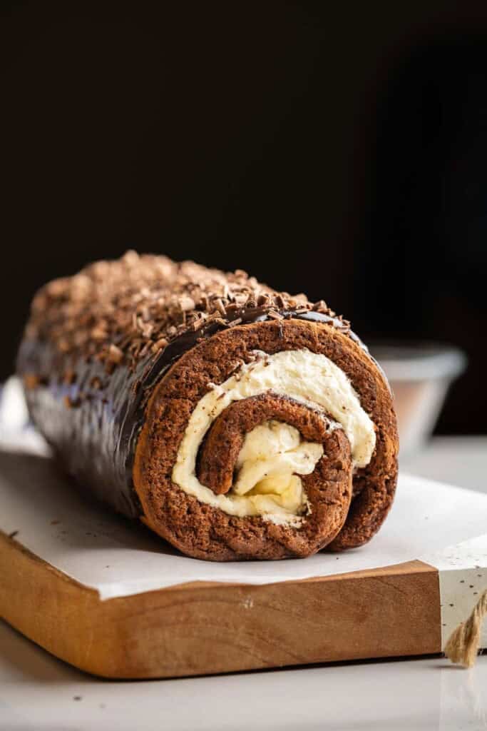 Gluten free chocolate swiss roll with a dark chocolate ganache and chocolate shavings on top placed on a cutting board.