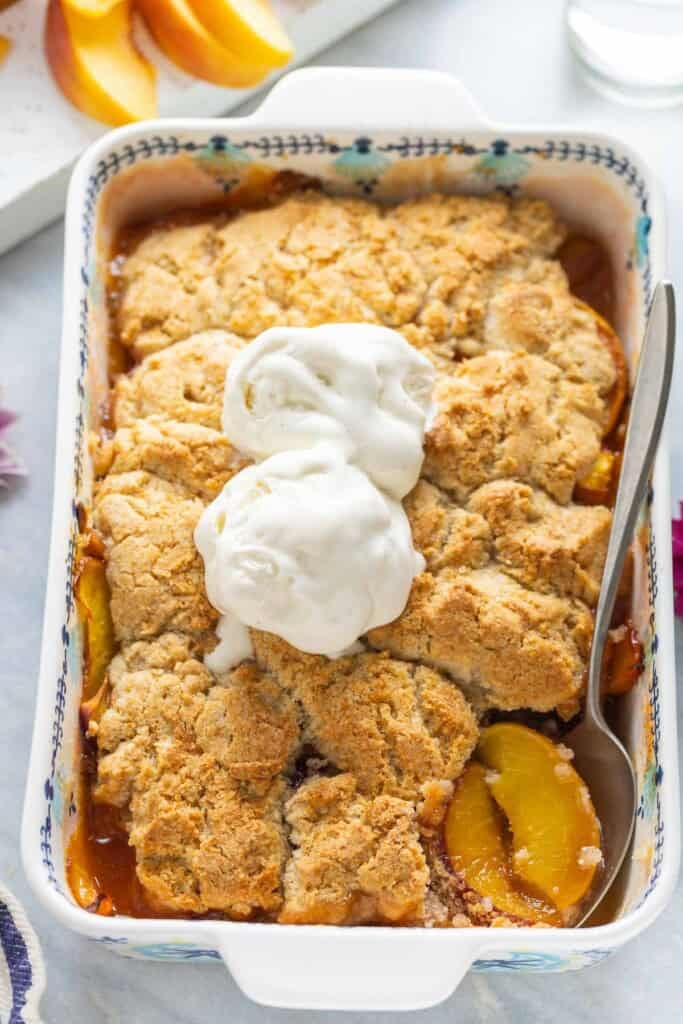 Gluten free peach cobbler in a baking dish with a couple scoops of vanilla ice cream on top