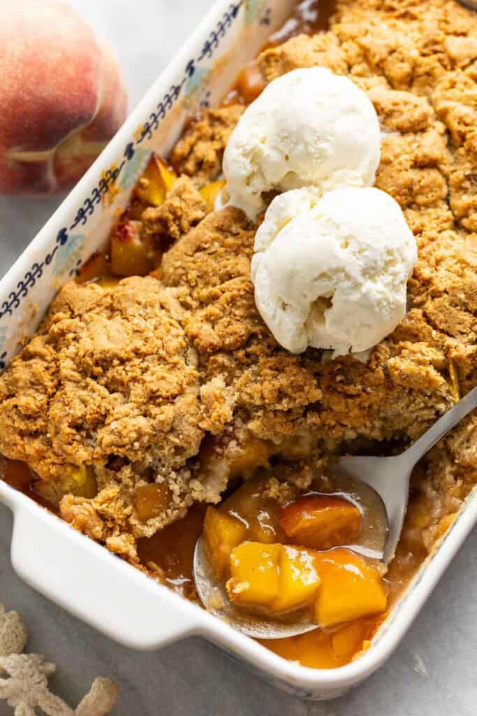 Gluten free peach cobbler with a couple scoops of vanilla ice cream on top and a serving spoon inside