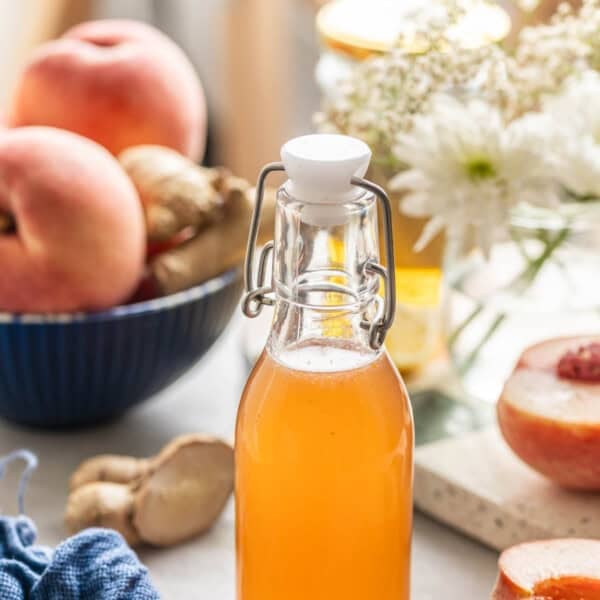 Homemade cold infused peach syrup in a small bottle with a swing top lid. Fresh peaches and ginger on the side.