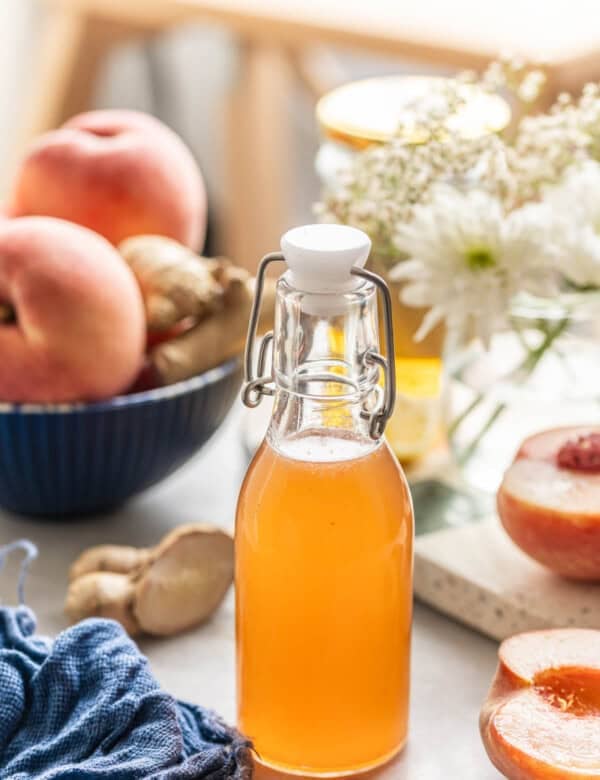 Homemade cold infused peach syrup in a small bottle with a swing top lid. Fresh peaches and ginger on the side.