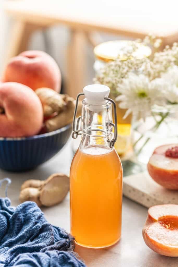 Homemade cold infused peach syrup in a small bottle with a swing top lid. Fresh peaches and ginger on the side.