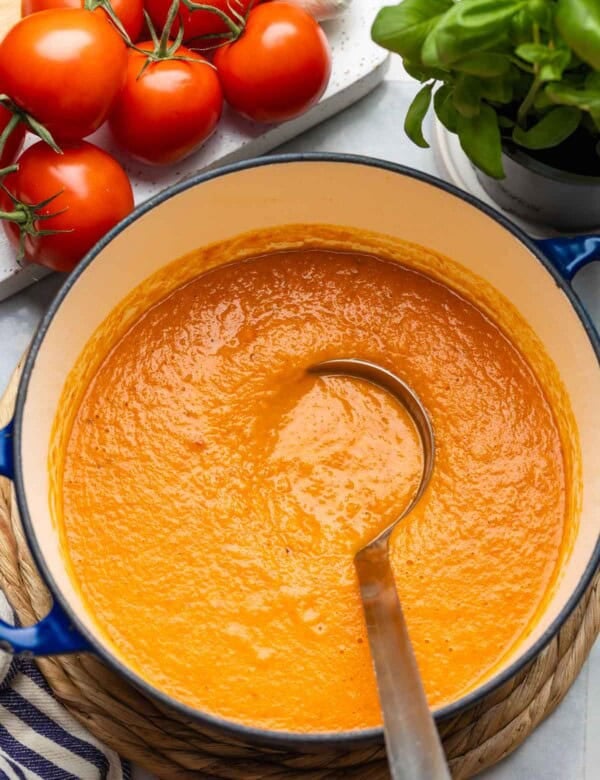 A large pot full of roasted tomato soup with a soup ladle inside, fresh tomatoes, garlic and basil on the side.