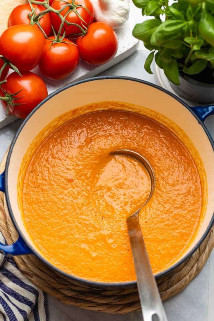 A large pot full of roasted tomato soup with a soup ladle inside, fresh tomatoes, garlic and basil on the side.