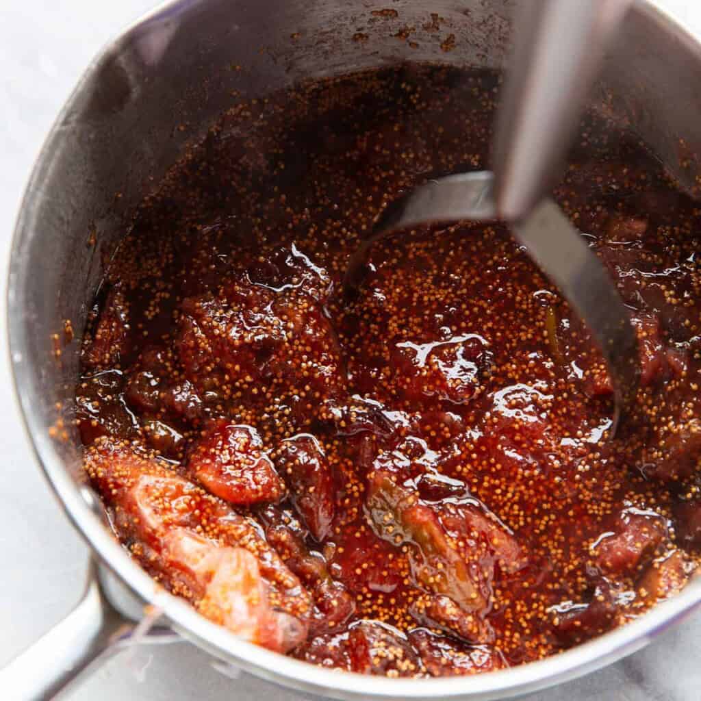 A sauce pan full of homemade fig jam with a potato mashed mashing up the larger pieces of fruit.