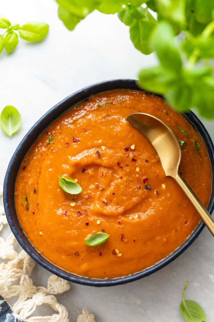 A bowl of roasted tomato soup with fresh basil leaves and red chili flakes sprinkles on top