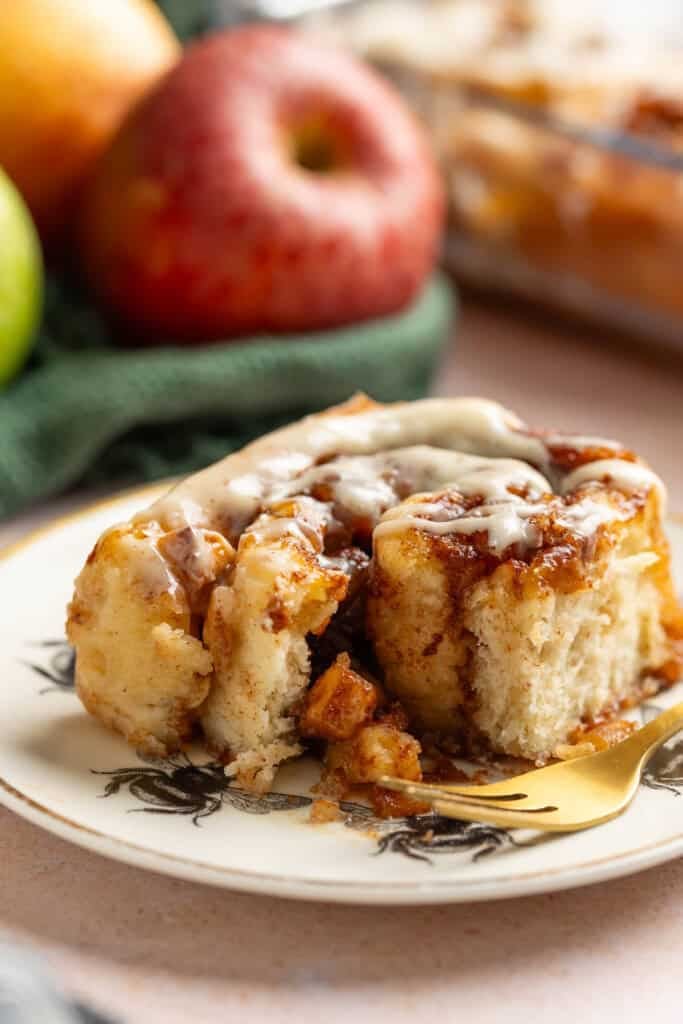 A small plate with a gluten free apple cinnamon roll with an apple pie filling and brown butter cream cheese frosting with a bite taken out.