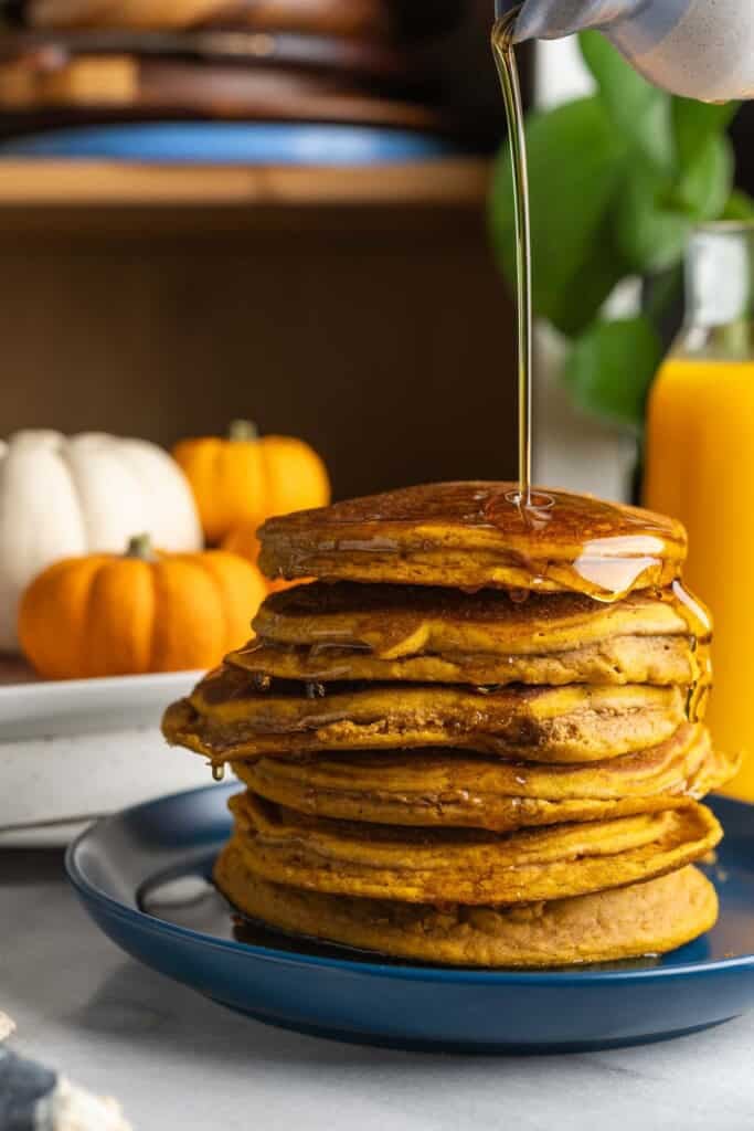 A stack of six gluten free pumpkin pancakes on a plate with maple syrup pouring over the top.
