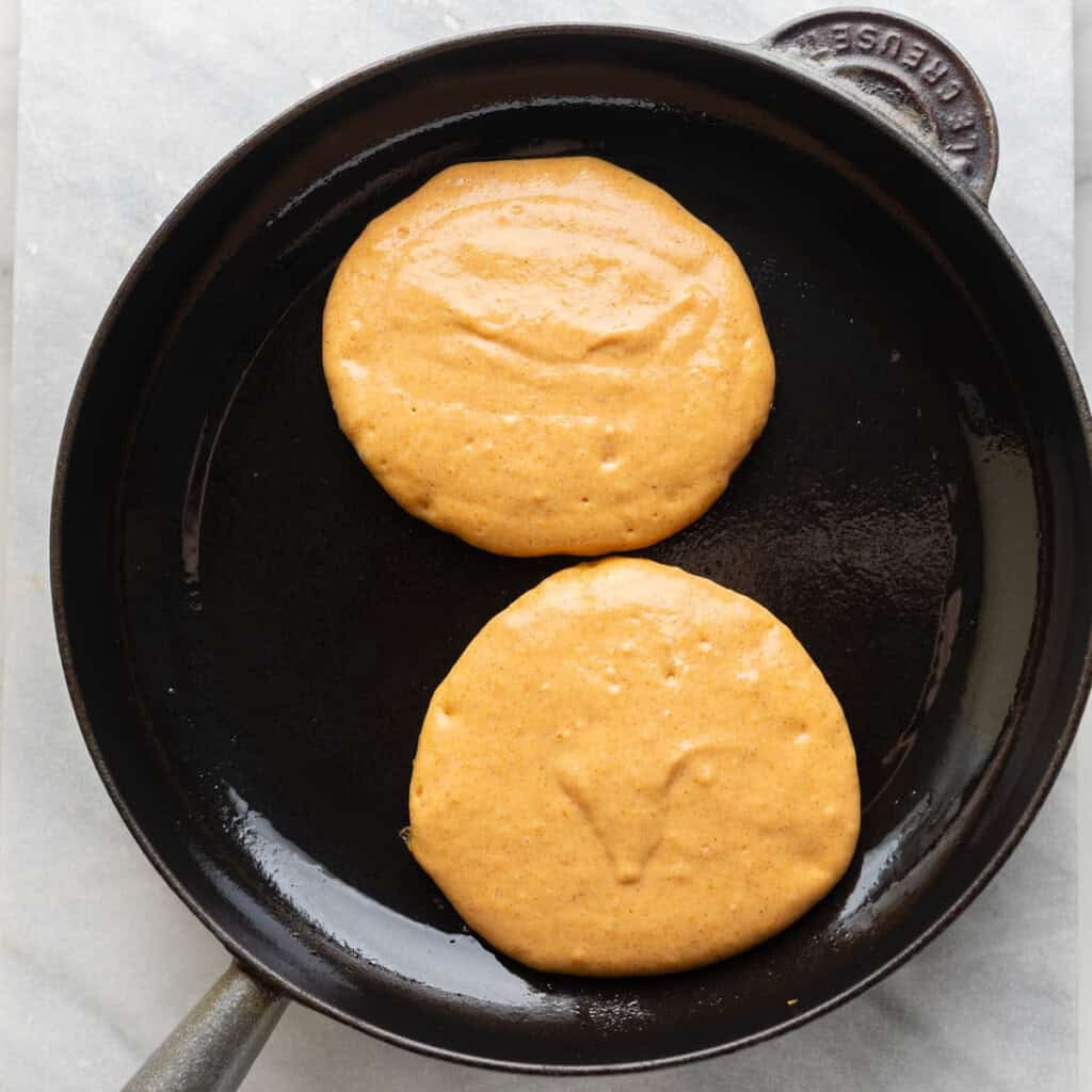 A cast iron skillet with 2 gf pumpkin pancakes cooking - the top side still uncooked.